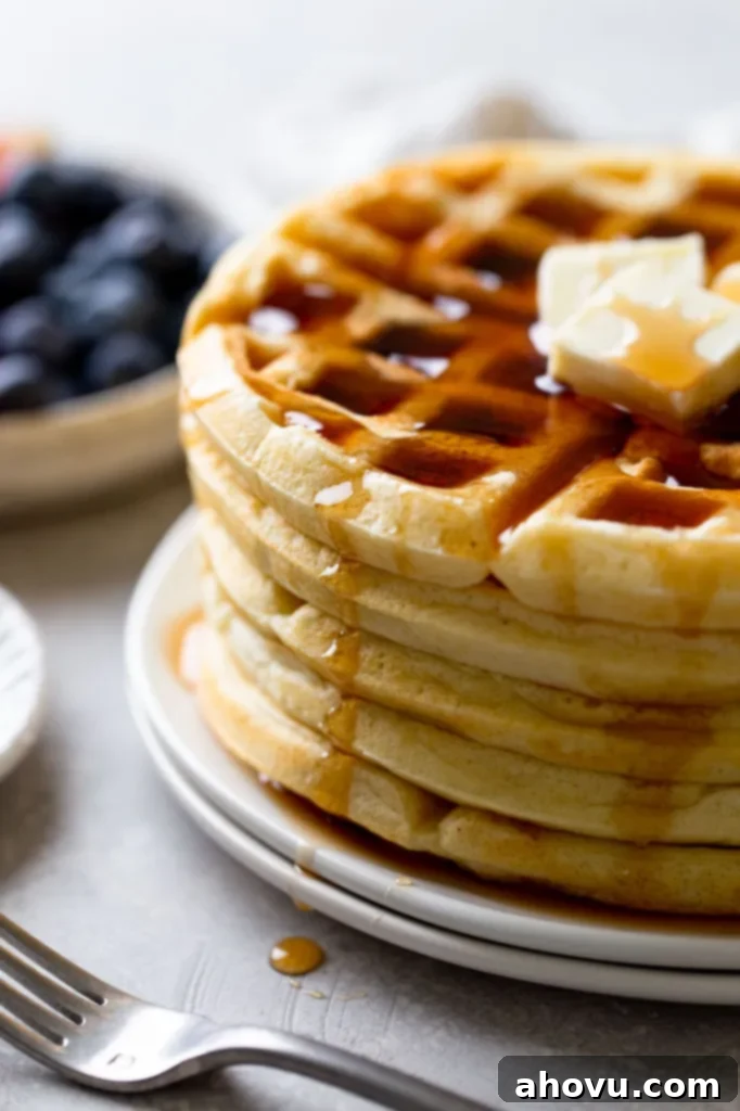 A side view of a stack of fluffy waffles topped with butter and syrup. A bowl of fresh berries rests in the background. 