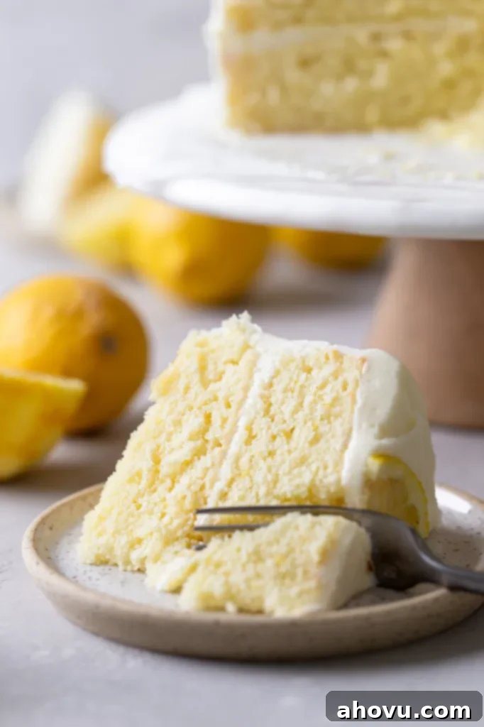 A slice of homemade lemon cake lying on its side on a speckled plate. A fork is digging into the slice. The rest of the cake is on a cake stand in the background. 