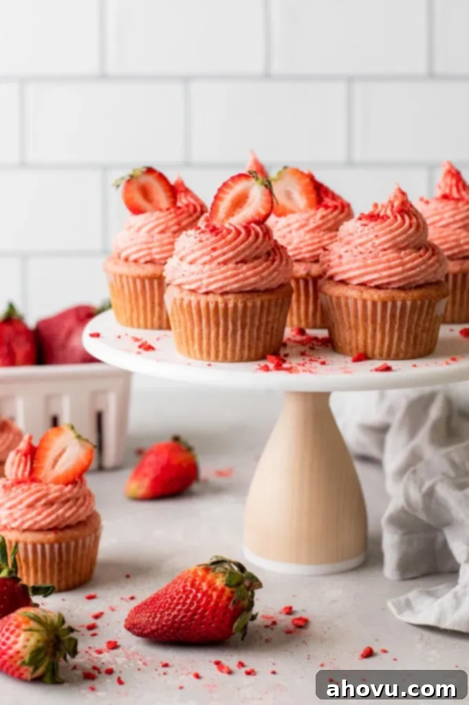 A side view of fresh strawberry cupcakes on a cake stand, elegantly frosted and garnished. Additional cupcakes and fresh berries are artfully arranged on the counter, hinting at the deliciousness within.