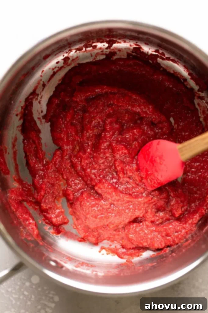 An overhead shot of vibrant red, reduced strawberry puree in a saucepan, with a rubber spatula resting in the pan, indicating its readiness for the next step in the recipe.