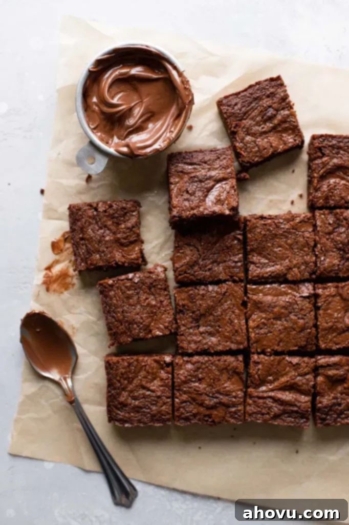 An overhead view of perfectly sliced Nutella brownies arranged on a square of parchment paper, with a small dish of Nutella and a spoon nearby, highlighting their rich, fudgy texture.