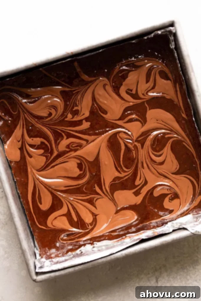 An overhead view of luscious brownie batter generously swirled with extra Nutella in a gleaming metal baking dish, ready for baking.