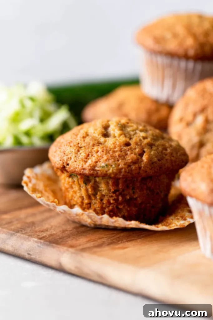 A close up of a zucchini muffin with the muffin liner pulled down. It's on a wood cutting board with more muffins surrounding it. 