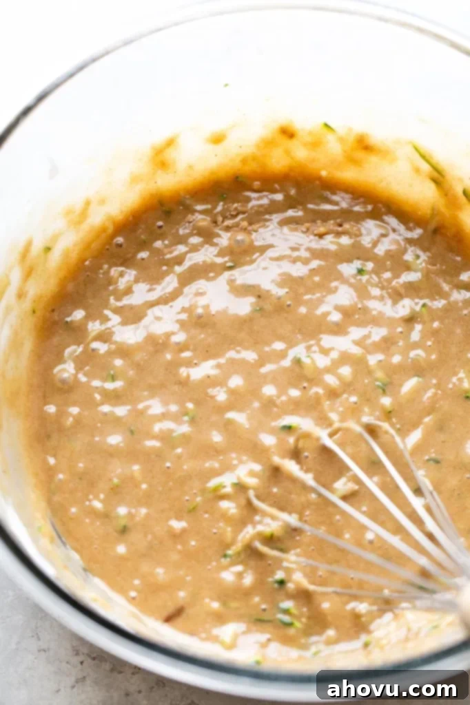 An overhead view of zucchini muffin batter in a glass mixing bowl with a whisk. 
