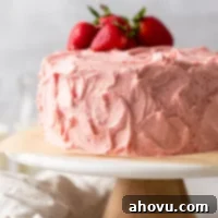 A strawberry cake with strawberry buttercream frosting sitting on top of a marble cake stand. Several fresh strawberries rest on top of the cake.