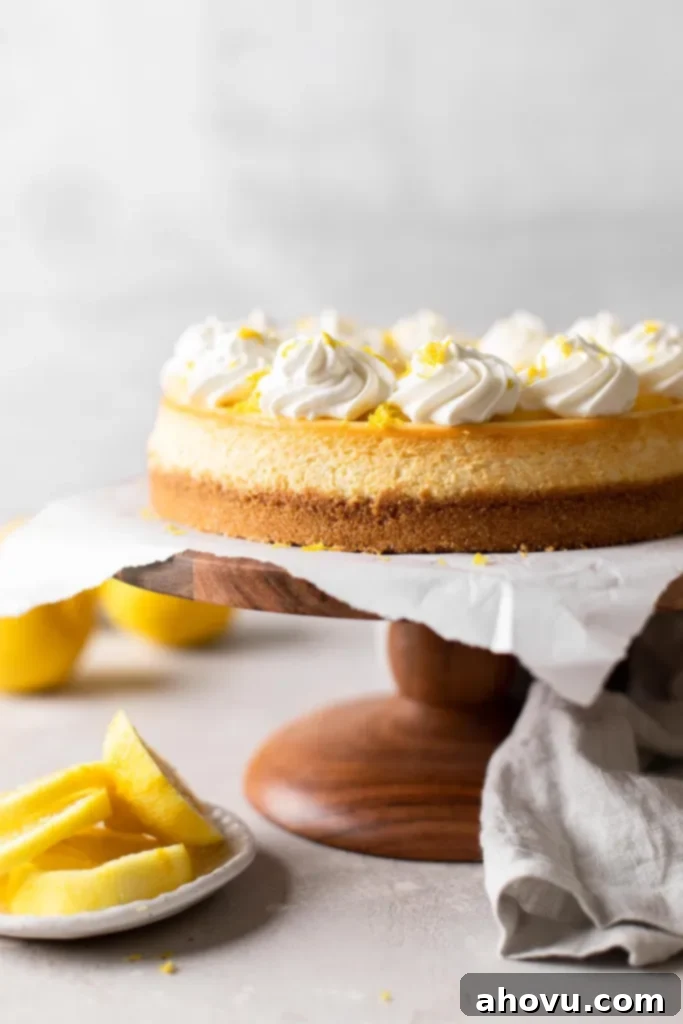A side view of a lemon cheesecake on a wooden cake stand. Lemon slices rest on a small plate nearby. 