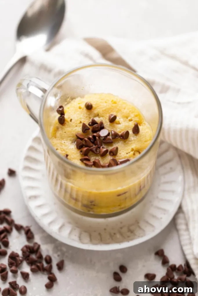 Decadent Chocolate Chip Mug Fix 39 A perfectly baked chocolate chip cookie in a ceramic mug, resting on a white dessert plate. The top is generously sprinkled with mini chocolate chips, and a spoon is visible in the soft-focus background, suggesting immediate enjoyment.