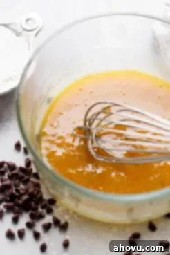 A glass mixing bowl holds a perfectly combined mixture of melted butter and sugars, with a whisk resting inside. This illustrates the first step in creating the delicious chocolate chip mug cookie dough.