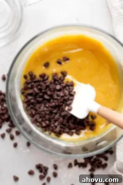 An overhead view showing mini chocolate chips being gently folded into the rich, raw cookie dough mixture with a rubber spatula, creating a perfectly blended batter for the microwave mug cookie.