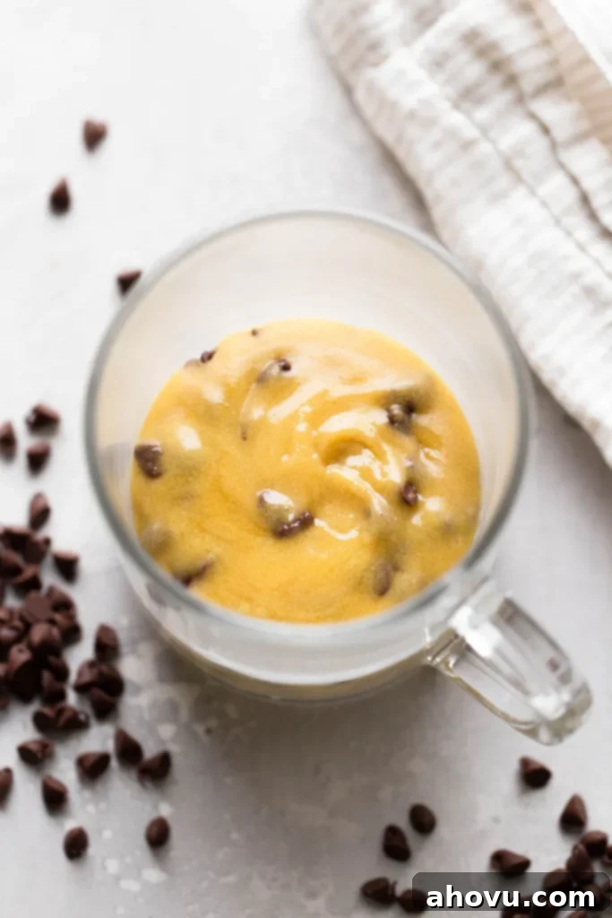 Decadent Chocolate Chip Mug Fix 4 An overhead view of rich, unbaked chocolate chip cookie dough neatly placed inside a clear glass mug, ready for the microwave. Mini chocolate chips are visible both in the dough and scattered around the base of the mug, hinting at the treat to come.