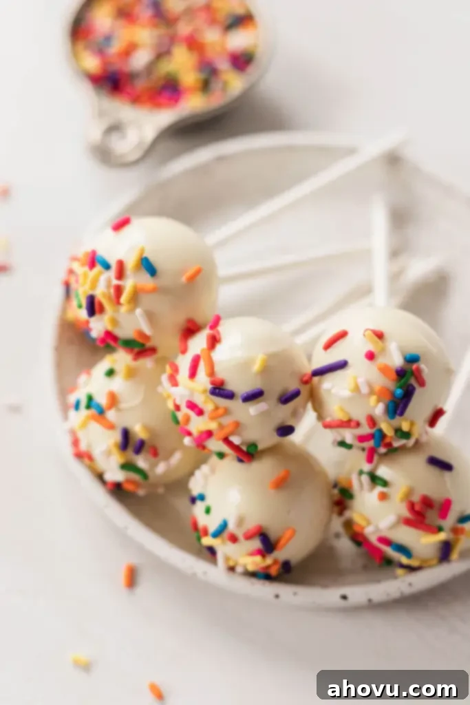 Six birthday cake pops resting on top of each other on a small white plate. Sprinkles in a measuring cup rest in the background. 