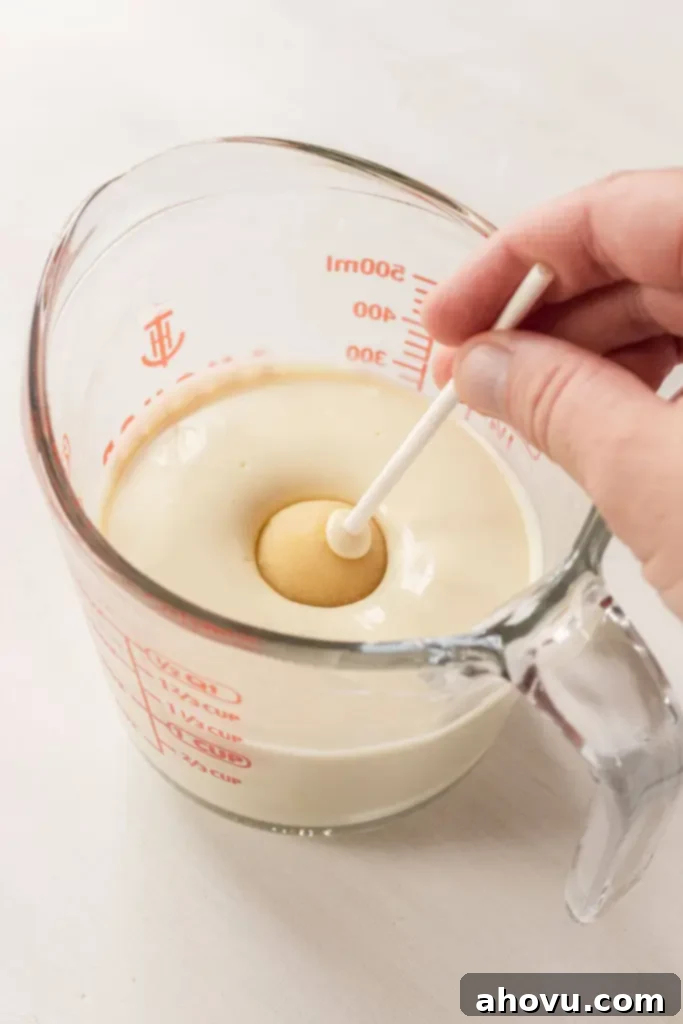 A vanilla cake pop being dunked into a glass of melted white chocolate. 