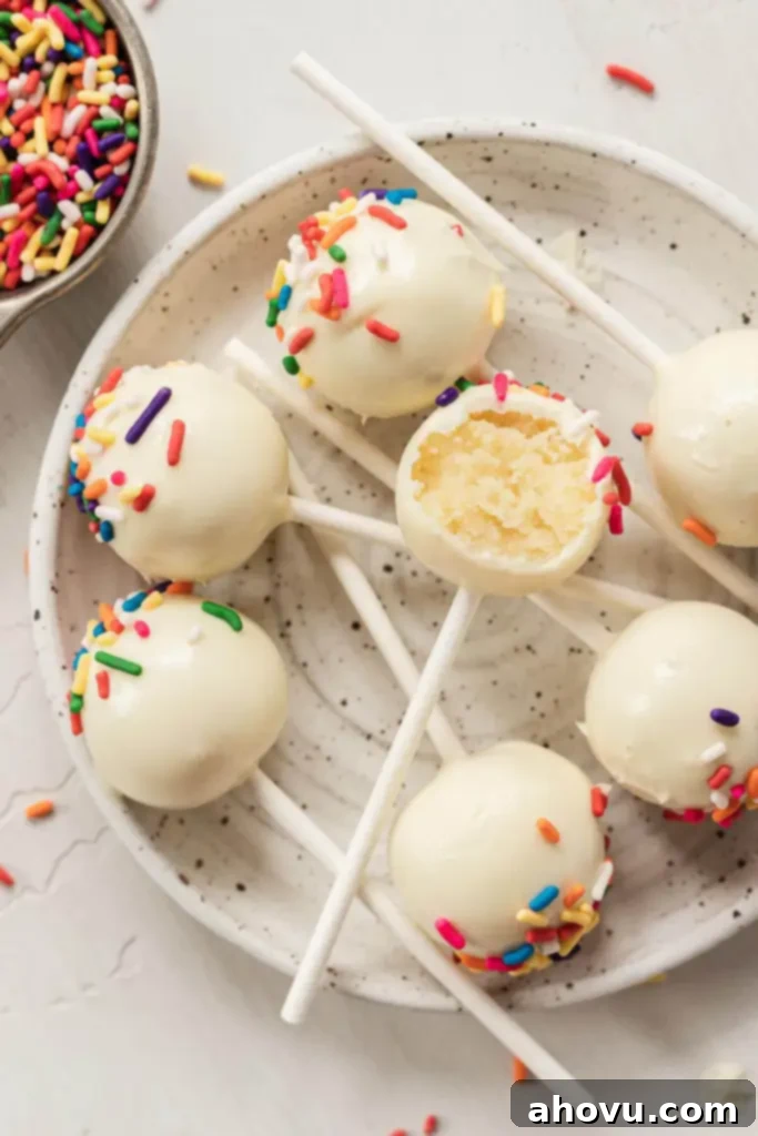 An overhead view of birthday cake pops on a small speckled plate. One cake pop has a bite missing. 
