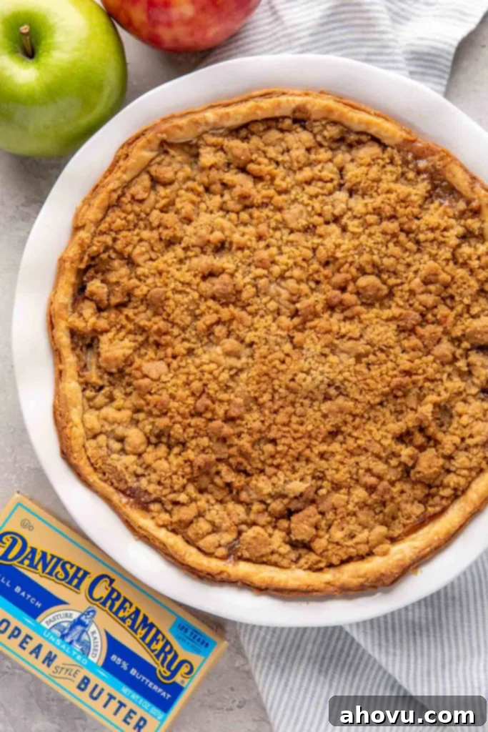 An apple crumble pie in a white pie dish. Two apples and a box of butter surround the pie dish.