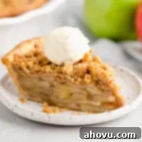 A slice of apple crumble pie topped with a scoop of ice cream on a white plate. Two apples and the rest of the pie are in the background.