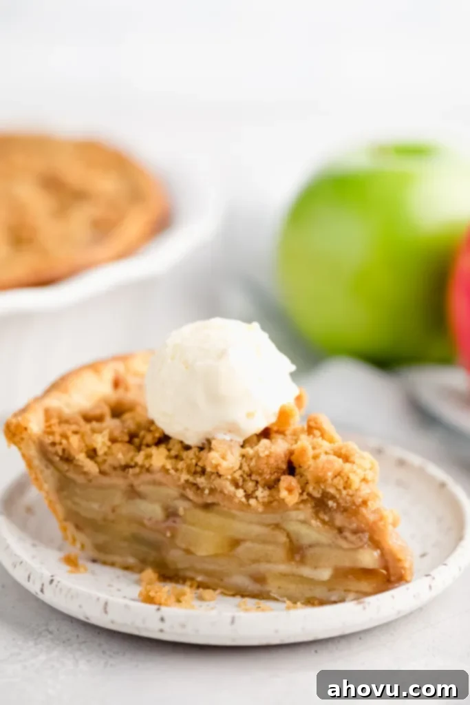 A slice of apple crumble pie topped with a scoop of ice cream on a white plate. Two apples and the rest of the pie are in the background.
