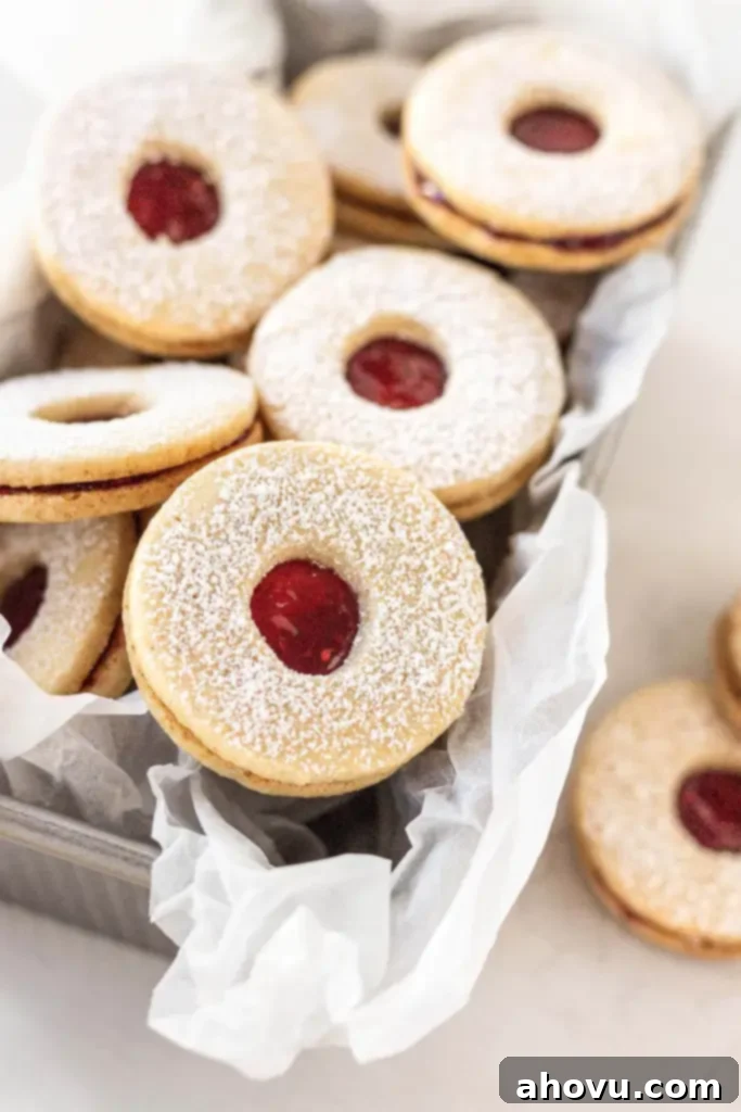 Peekaboo Jam Cookies 6 A charming close-up of classic Linzer cookies, artfully piled in a parchment paper-lined loaf pan, dusted with confectioners' sugar and ready to be served or gifted.