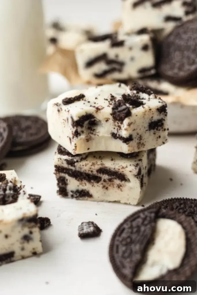 Oreo Cookies and Cream Fudge 7 Two pieces of smooth, no-bake Oreo fudge elegantly stacked, highlighting their thick, creamy texture and visible cookie chunks. Additional fudge slices and whole Oreo cookies are artfully arranged around the stack, creating a tempting display.