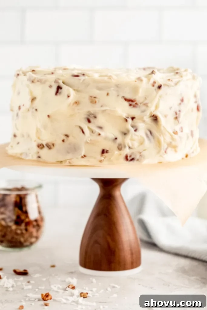 An Italian Cream Cake on top of a wooden cake stand with a marble top. A jar of chopped pecans and two milk jugs rest in the background.