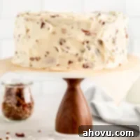 An Italian cream cake on a wooden cake stand with a marble top. A jar of chopped pecans and two milk jugs rest in the background.