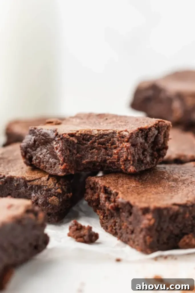 A close-up view of a rich, fudgy cocoa powder brownie with a corner bite missing, perfectly stacked atop two additional brownies on a white plate, highlighting its moist interior. 