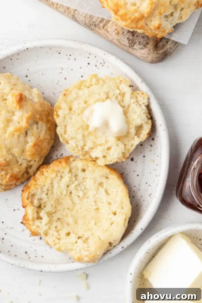 No-Fuss Fluffy Biscuits 7 An overhead view of a fresh batch of homemade drop biscuits on a white plate. One biscuit is halved and spread with butter, highlighting its soft interior.