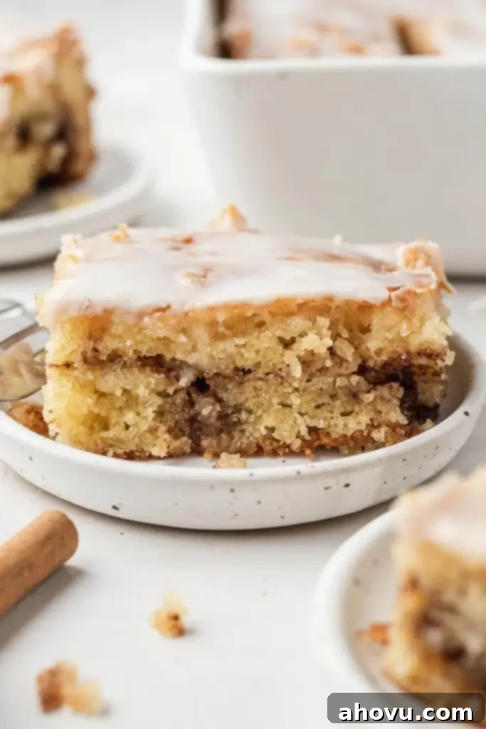 Glazed Cinnamon Honey Bun Cake 2 A slice of iced honey bun cake on a speckled white dessert dish. Additional cake slices rest in the foreground and background.