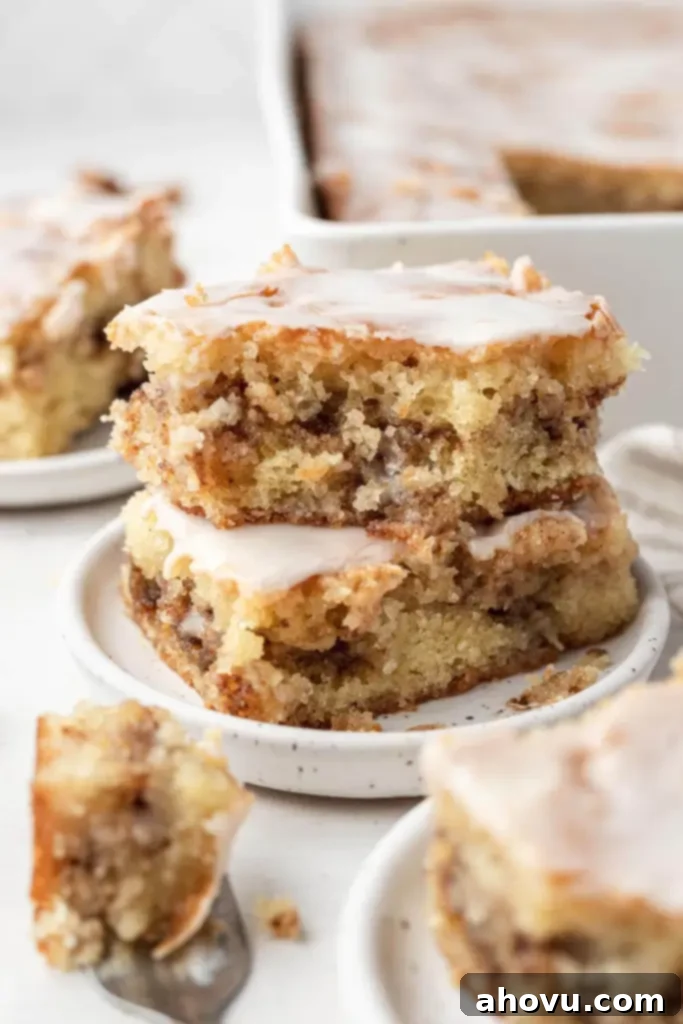 Glazed Cinnamon Honey Bun Cake 6 Two slices of honey bun cake, stacked on a white speckled dessert plate. Additional cake slices and a baking pan rest in the foreground and background.