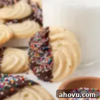 Golden Butter Delights 8 A butter cookie that has been dipped in chocolate sitting up against a glass of milk. More cookies and a bowl of sprinkles are around it.
