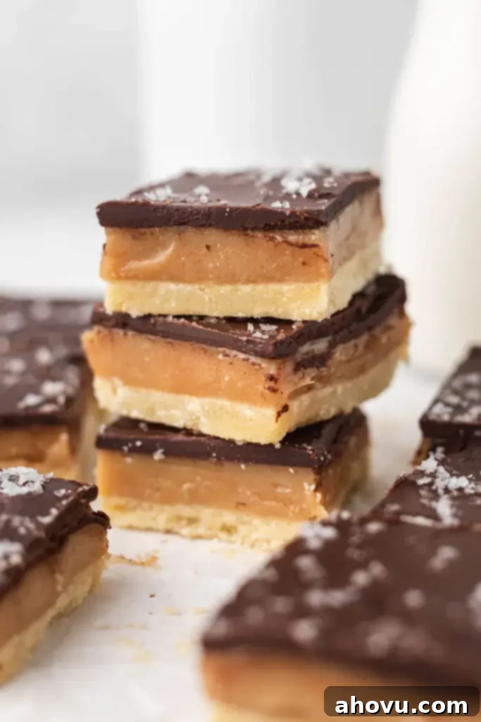 Millionaire Shortbread 2 A stack of three millionaire shortbread bars, expertly cut and topped with flaky sea salt, surrounded by additional bars on parchment paper.