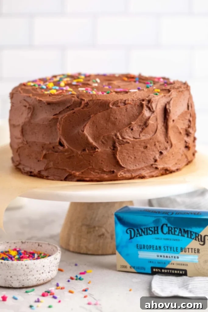 A beautifully frosted yellow cake sitting on top of a marble cake stand. A bowl of colorful sprinkles and a box of premium Danish Creamery butter are strategically placed next to the cake stand, highlighting key elements of the recipe.