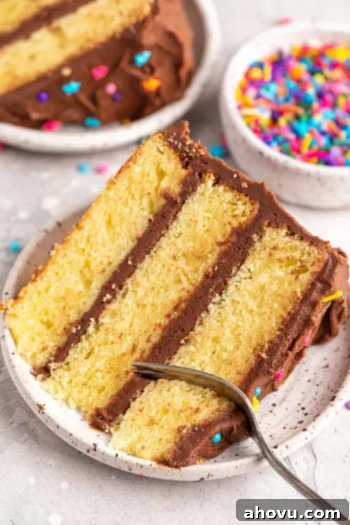 Two generous slices of yellow cake displayed on white speckled plates. One slice is being delicately cut with a fork, emphasizing the cake's tender and moist texture. The background shows the main cake on a stand.