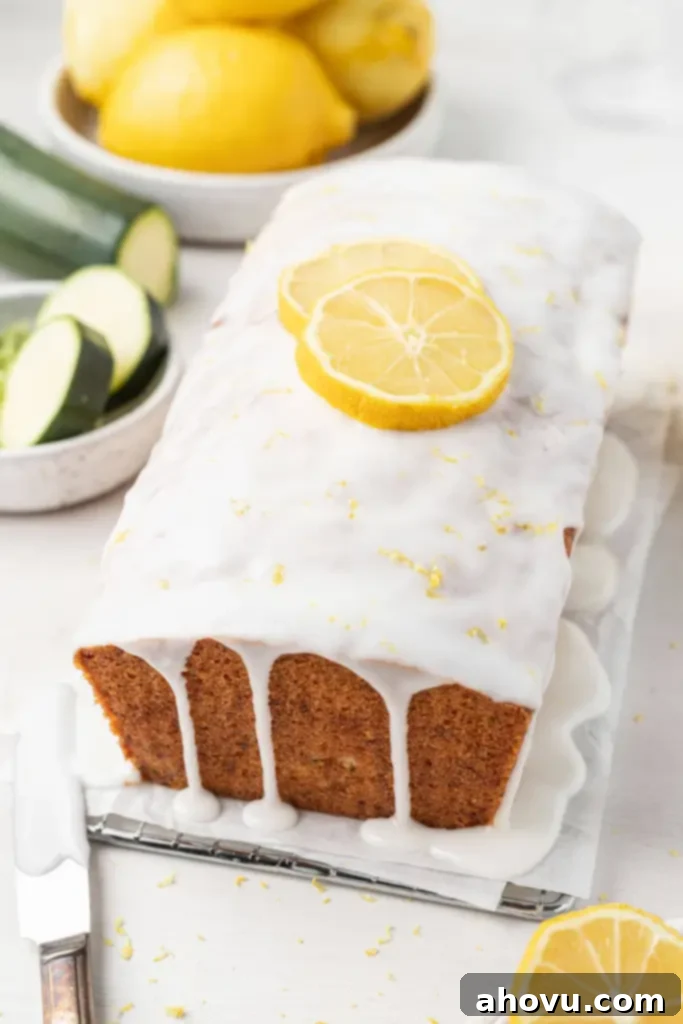 A loaf of glazed lemon zucchini bread on a parchment-lined wire rack. Dishes of sliced zucchini and lemons are in the background. 