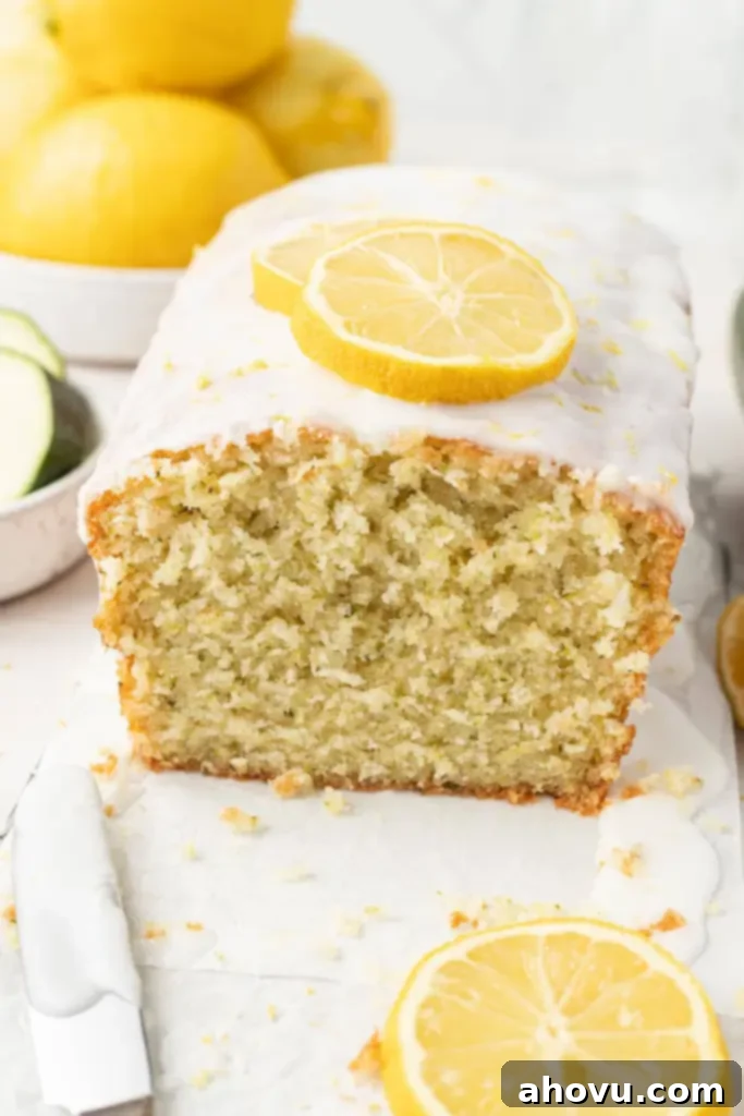 A loaf of glazed lemon zucchini bread, with a slice missing. A bowl of lemons rests in the background. 