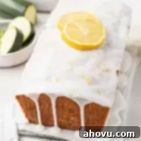 A loaf of lemon zucchini bread topped with two lemon slices. Some sliced zucchini rests in a small bowl on the side.