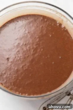 An overhead view of rich, smooth chocolate cake batter in a clear glass mixing bowl, perfectly mixed and ready for baking into delicious cake layers.