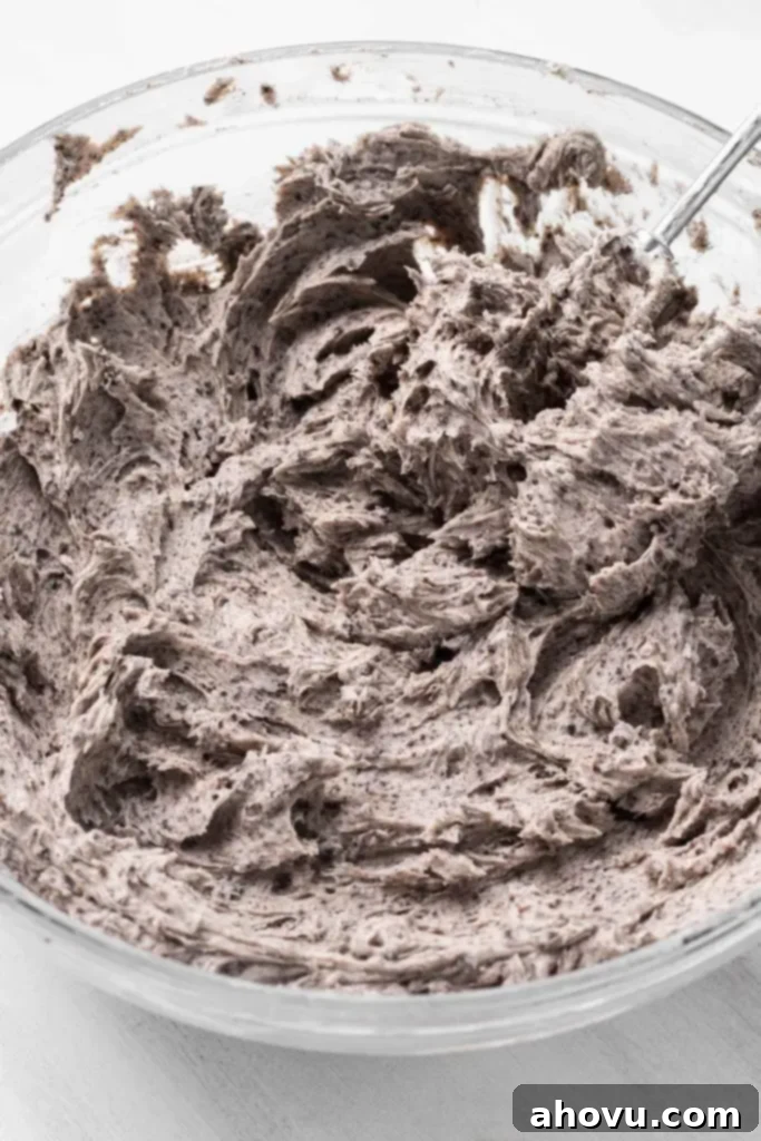Decadent Oreo Cake 4 A large glass mixing bowl filled with creamy, delicious Oreo buttercream frosting, showcasing visible flecks of crushed Oreos throughout. The frosting is perfectly smooth and ready for cake decorating.