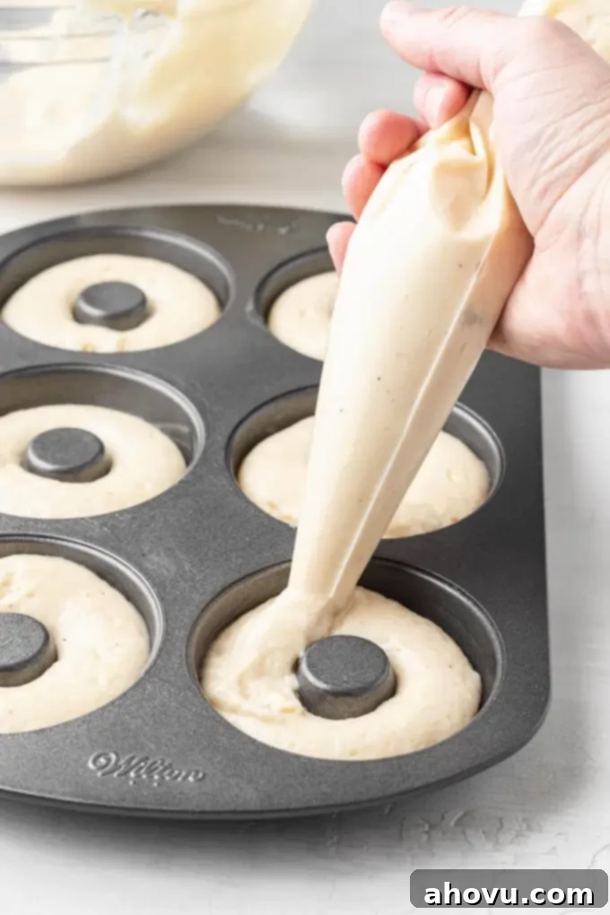Baked donut batter being expertly piped from a pastry bag into the cavities of a donut pan, ensuring even filling.