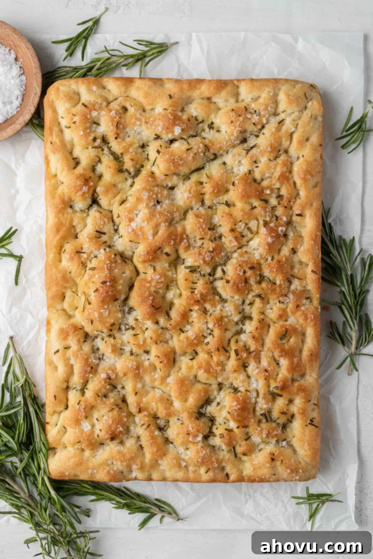 An overhead view of a loaf of baked focaccia, surrounded by fresh rosemary sprigs and a small bowl of sea salt. 