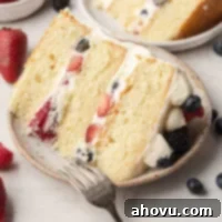 A slice of Chantilly cake on a speckle white plate. A fork rests on the plate and blueberries and strawberries are lying around it.