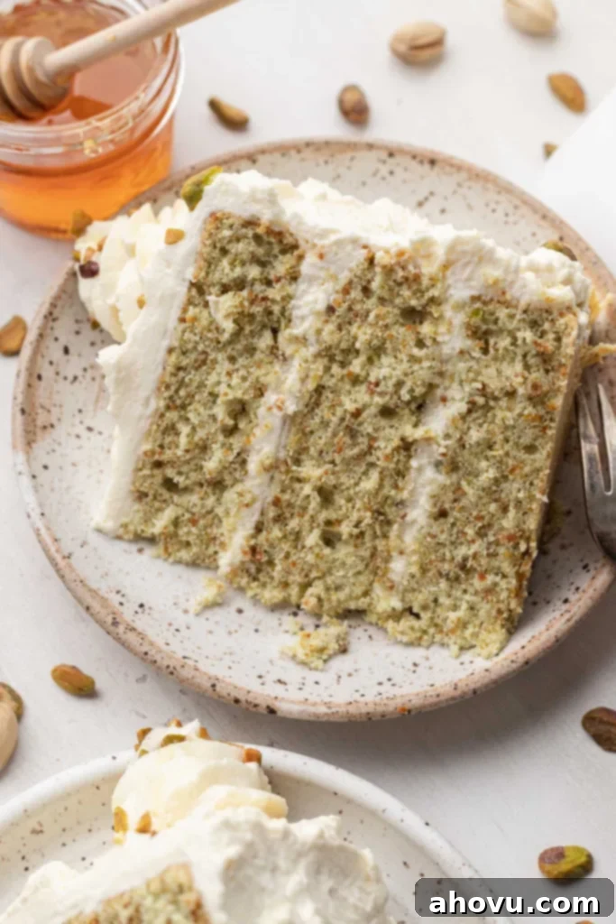 A slice of pistachio nut cake on a speckled white dessert plate, with honey on the side. 