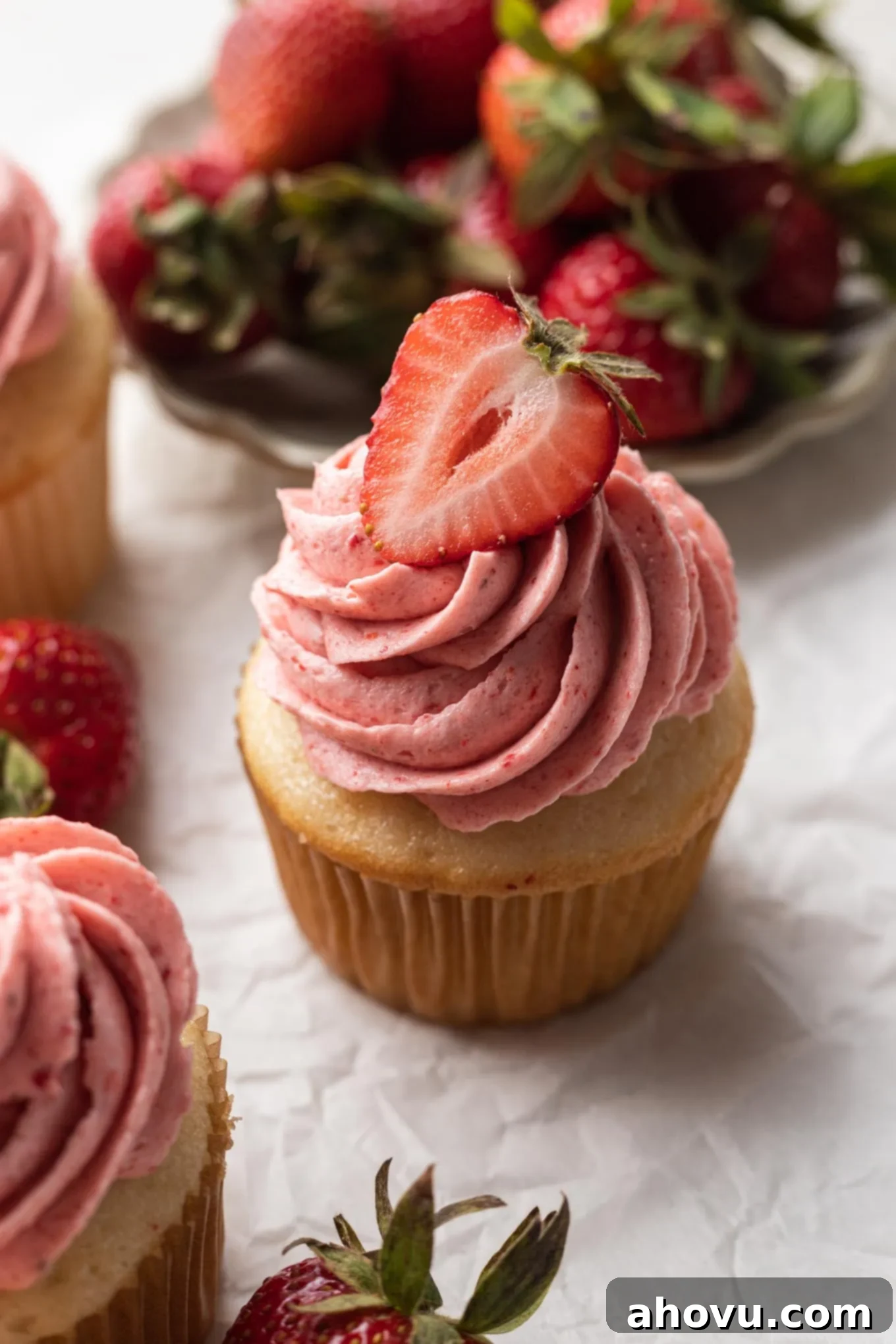 A vanilla cupcake topped with fluffy pink strawberry frosting and garnished with a fresh strawberry half, set against a bright background.