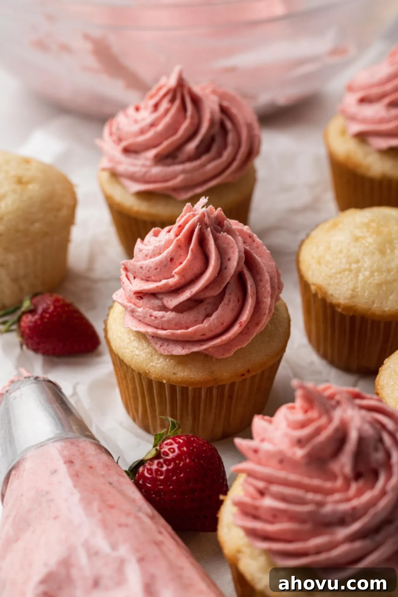 A charming close-up of several vanilla cupcakes, some frosted with delightful strawberry buttercream, and a piping bag overflowing with the same frosting in the foreground, ready for more decorating.