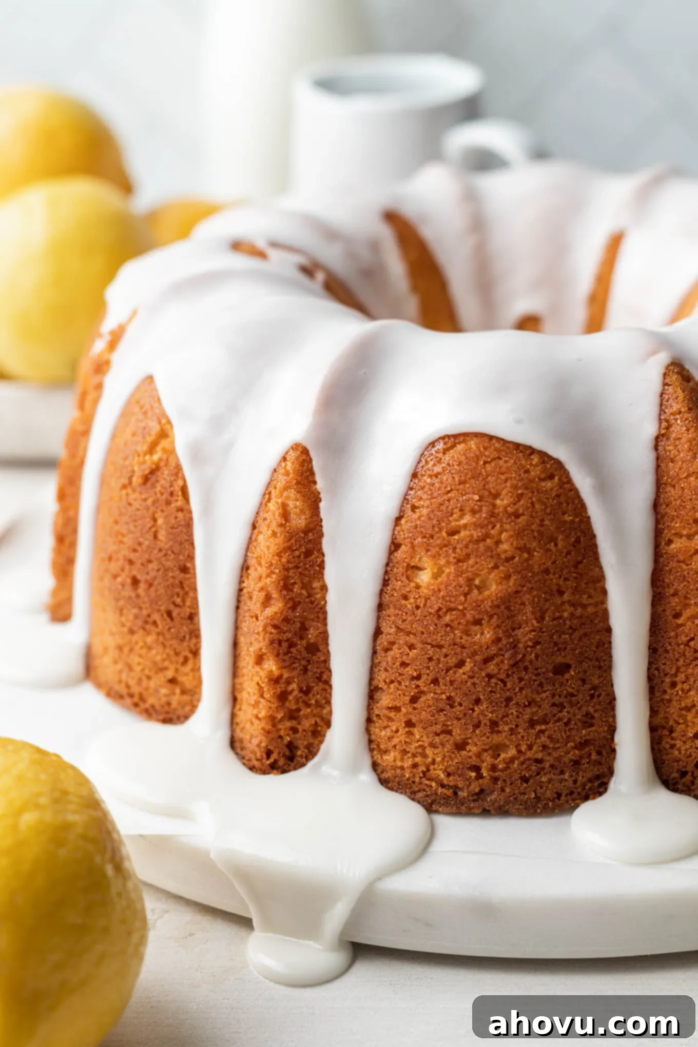 A glazed lemon pound cake on a white cake plate, with lemons in the foreground and background. 