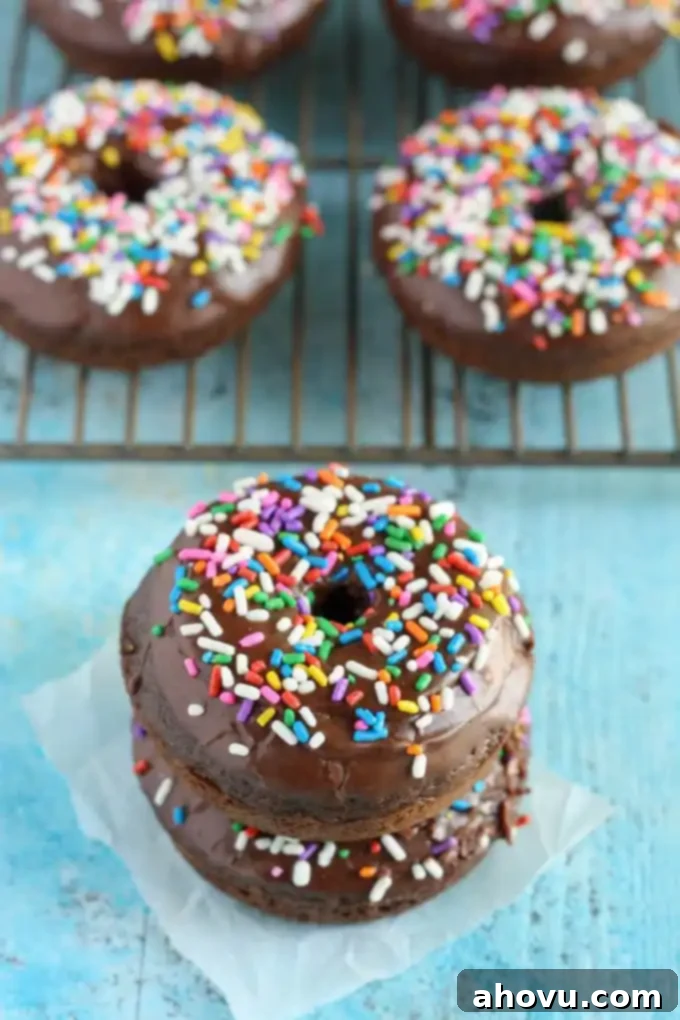 A stack of two frosted chocolate donuts on a square of parchment paper. More donuts are on a cooling rack in the background. 