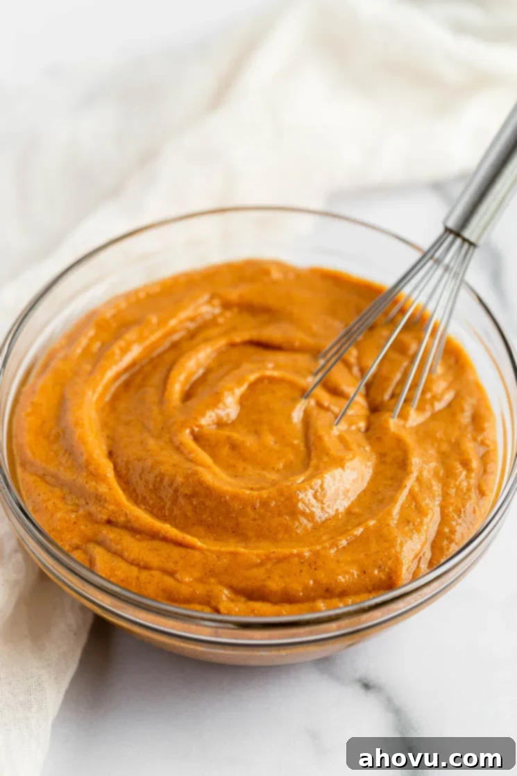 A clear glass mixing bowl filled with smooth, orange-hued pumpkin cupcake batter, with a silver metal whisk resting beside it.