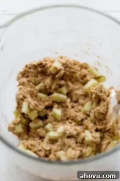 A glass mixing bowl holding golden cookie dough with finely chopped apples folded into it, ready for baking.