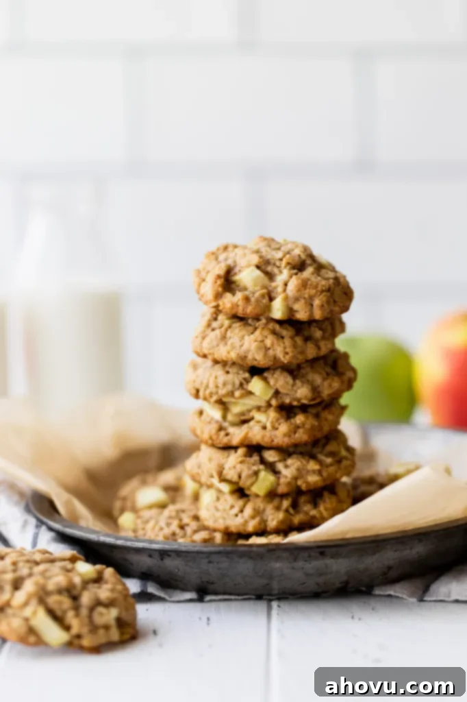 A stack of golden-brown apple oatmeal cookies resting in a rustic metal baking dish. A container of milk and a couple of fresh apples sit artfully in the background, suggesting a perfect snack or dessert.