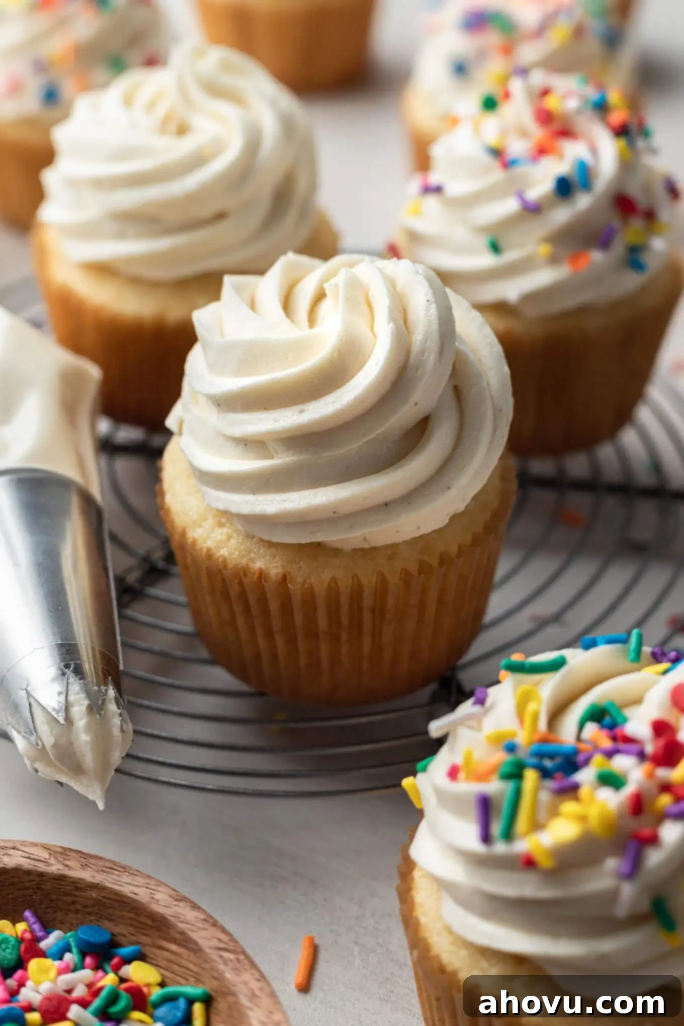 Frosted vanilla cupcakes next to a piping bag and dish of sprinkles. 