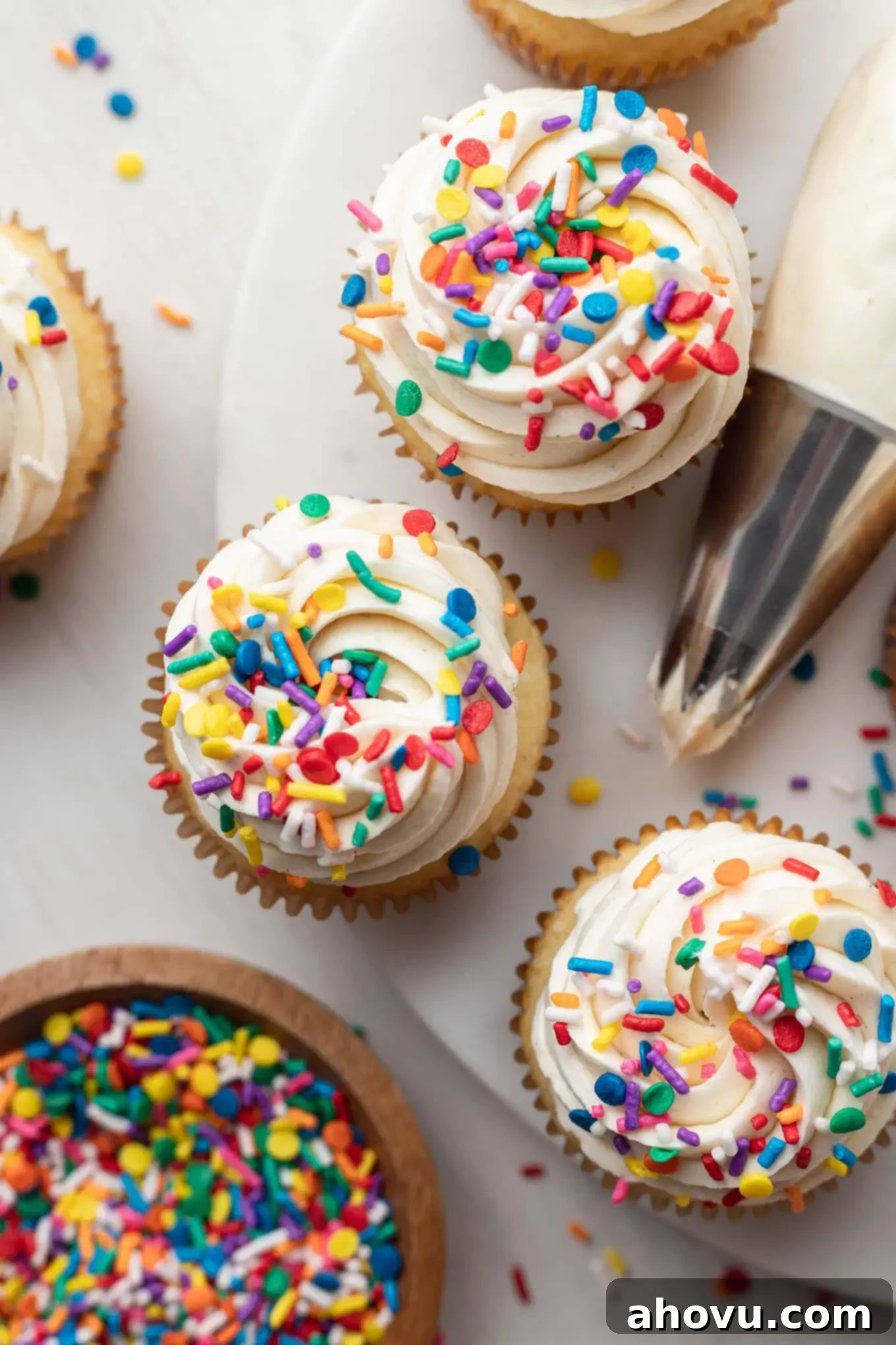 An overhead view of three cupcakes with white frosting next to a piping bag and sprinkles. 